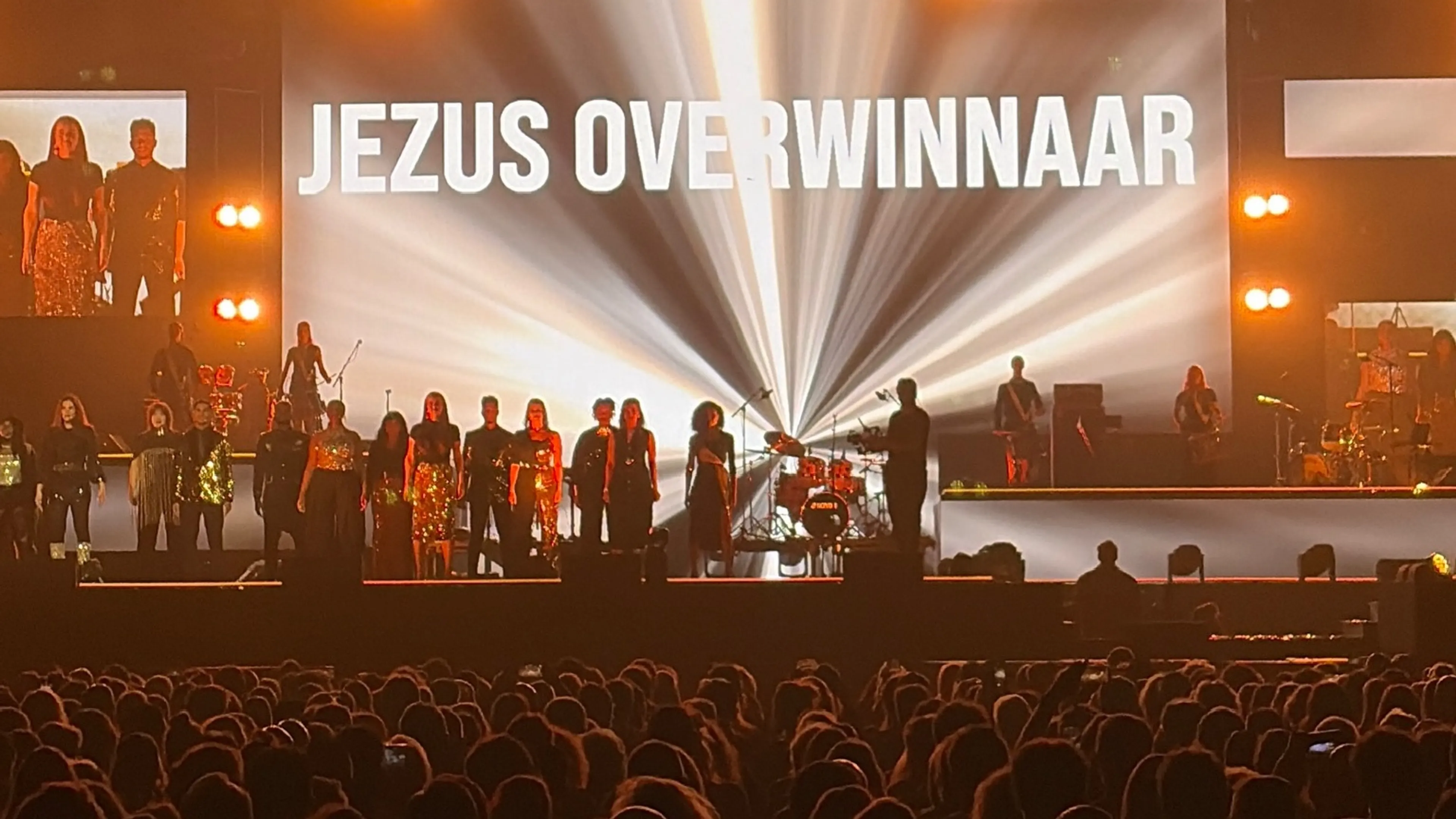 JEZUS OVERWINNAAR — een grootschalige liveperformance of worship-bijeenkomst met een grote groep performers op een podium, waarbij een enorme projectiescherm de tekst toont en felle lichtbundels de scène verlichten.