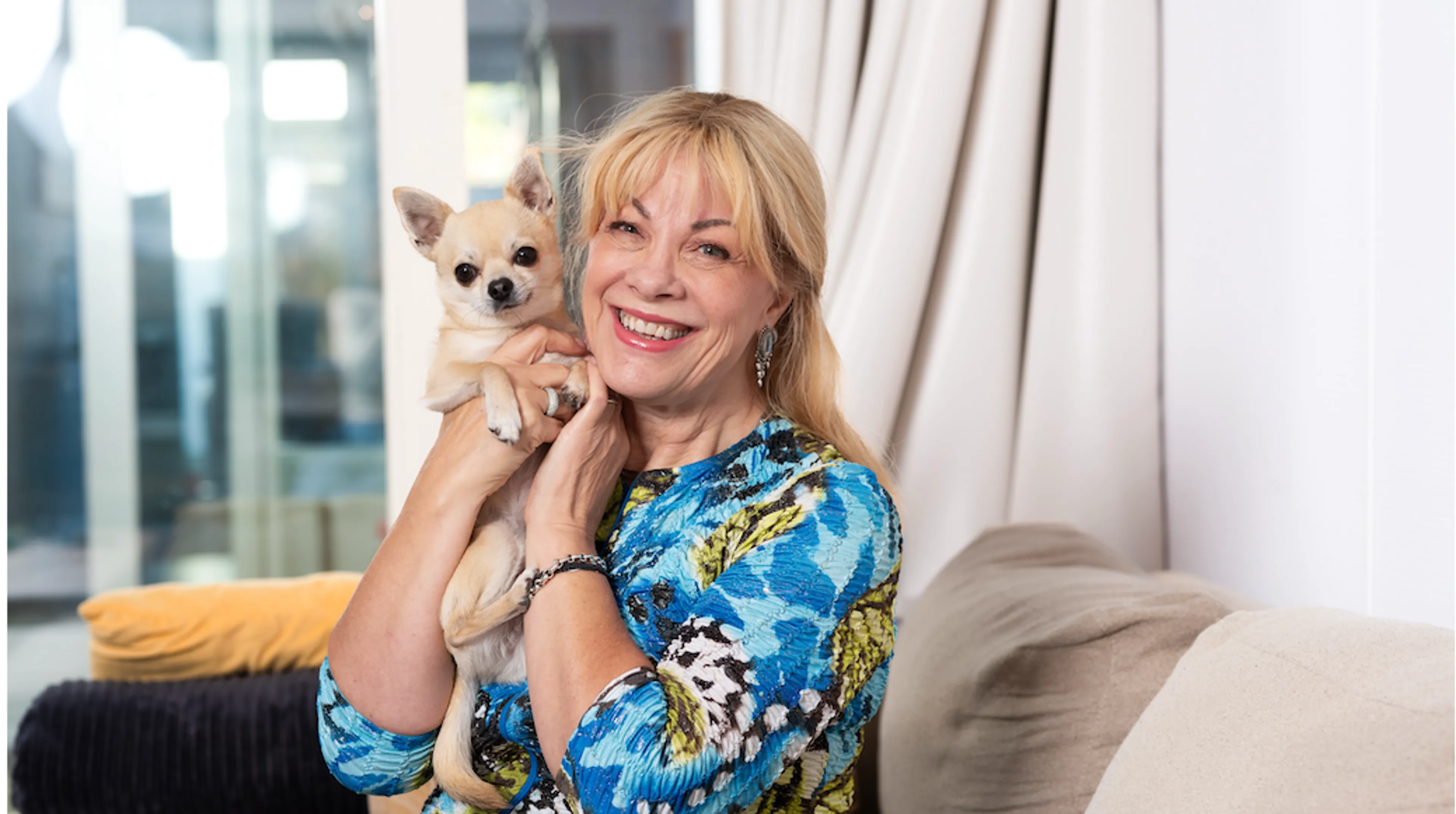 Op de foto zijn een middelbare tot oudere vrouw met blond haar en een brede glimlach en een kleine lichtbeige Chihuahua te zien, knuffelend tegen haar gezicht terwijl de hond recht in de camera kijkt. De vrouw draagt een opvallende blauw-groene top met een abstract bloemachtig patroon en zit bij een beige bank in een moderne woonkamer met grote ramen.
