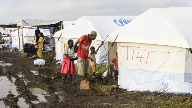 Een man en twee kinderen lopen door de modderige aarde over het tentenkamp met witte tenten.
